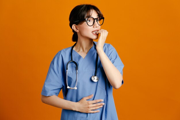 Tense grabbed chin young female doctor wearing uniform fith stethoscope isolated on orange background