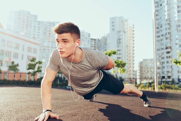 Tense handsome guy with a good body at workout in the morning on stadium. He wears sport clothes, listening to music through headphones,  push-ups with one hand.