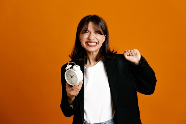 Tense holding alarm clock young beautiful female wearing black jacket isolated on orange background