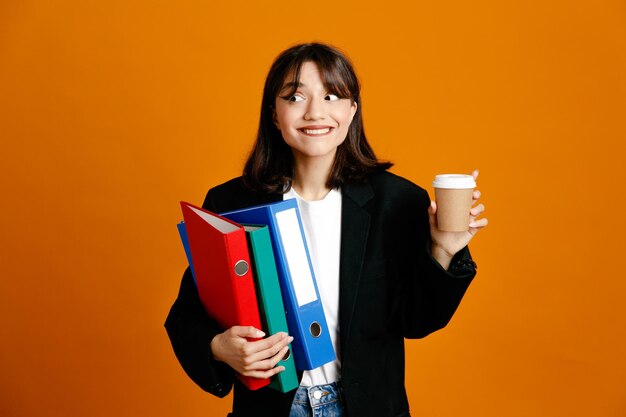 オレンジ色の背景で隔離の黒いジャケットを着ているコーヒーカップの若い美しい女性と緊密な保持フォルダー