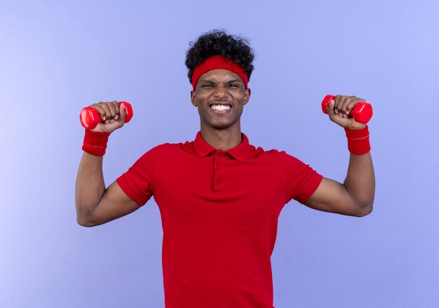 Tense young afro-american sporty man wearing headband and wristband raising dumbells isolated on blue wall