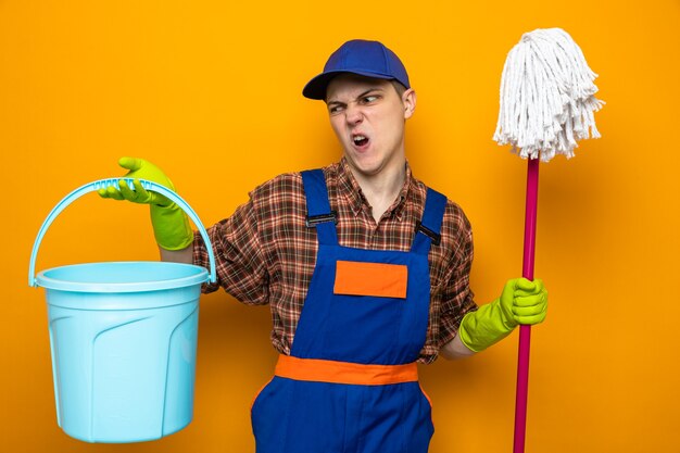 Tense young cleaning guy wearing uniform and cap with gloves holding mop and looking at bucket in his hand 