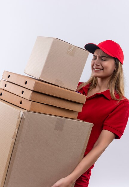 Tense young delivery girl wearing red uniform and cap holding many boxes isolated on white