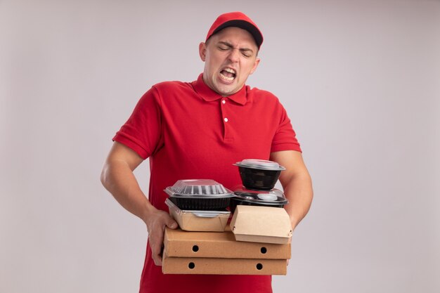 Tense young delivery man wearing uniform with cap holding food containers on pizza boxes isolated on white wall