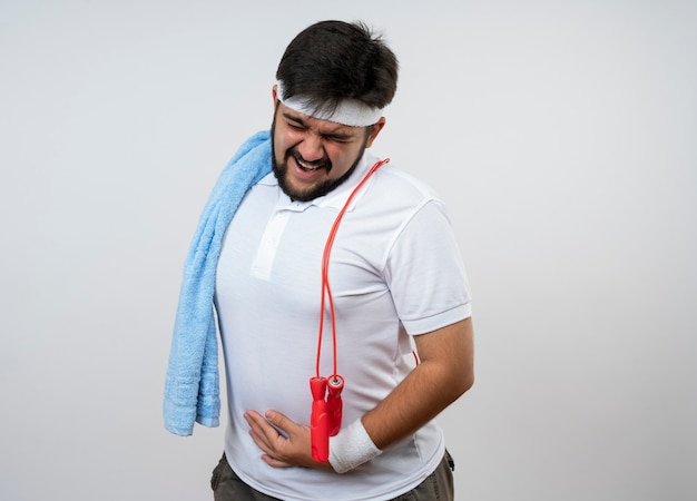 Tense young sporty man wearing headband and wristband with towel and jump rope on shoulder putting hand on stomach isolated on white wall