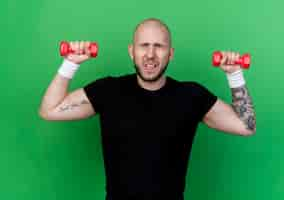 Free photo tense young sporty man wearing wristband exercising with dumbbells isolated on green