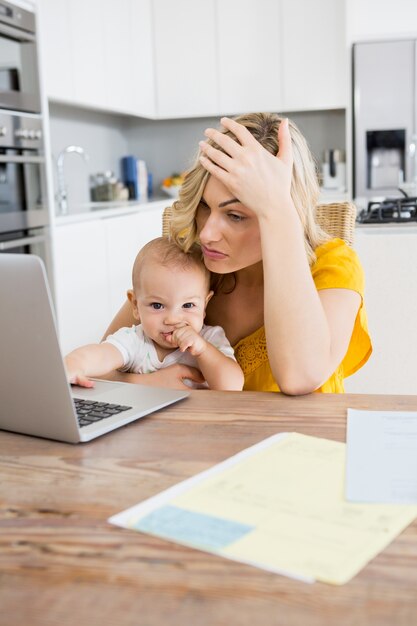 Tensed mother using laptop with her baby boy in kitchen