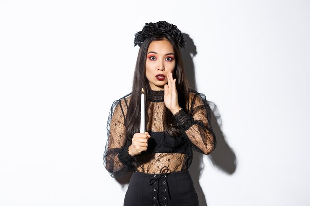 Tensed and worried asian woman in witch costume holding lit candle and whispering, standing over white background.