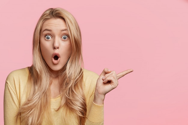 Бесплатное фото Испуганная молодая женщина предупреждает вас о чем-то, показывает указательным пальцем в сторону, держит широко открытым рот и смотрит на глаза, изолированно от розовой стены рекламная концепция