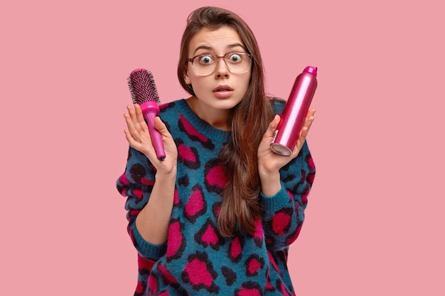 Free photo terriffied attractive lady in sweater and spectacles, makes hairdress with hairspray and comb