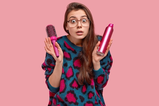 Terriffied attractive lady in sweater and spectacles, makes hairdress with hairspray and comb