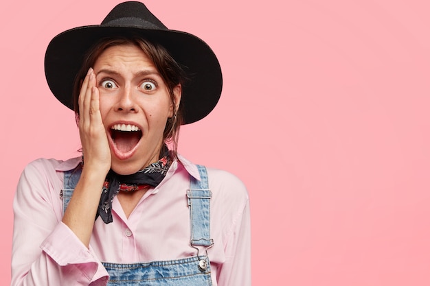 Free photo terrific cute female keeps hand on cheek, looks surprisingly, opens mouth widely, wears black hat and casual denim overalls, stands against pink wall