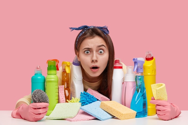 Terrific young woman with unexpected gaze, wears headband, has many detergents on table, shocked wth dirty room, wears protective gloves