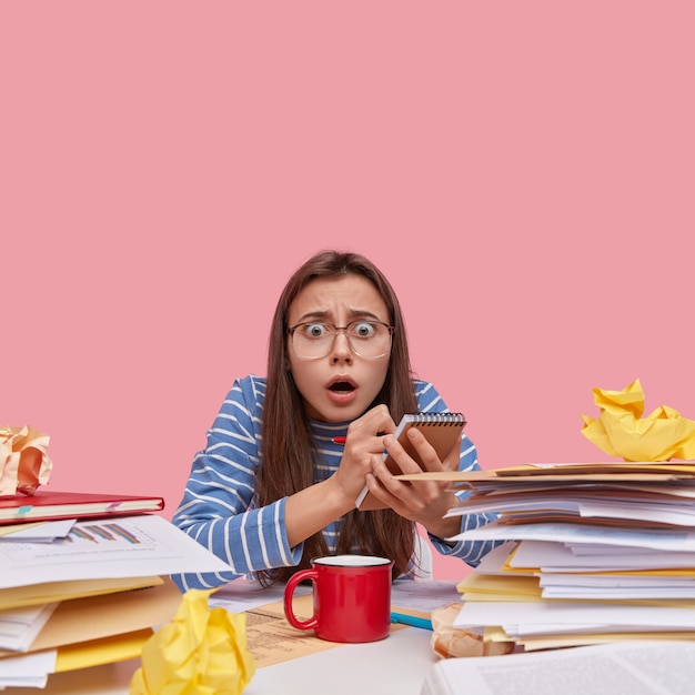 Free photo terrified beautiful lady has scared expression, wears big spectacles and striped clothes, makes notes in notepad, writes list to do, busy with work