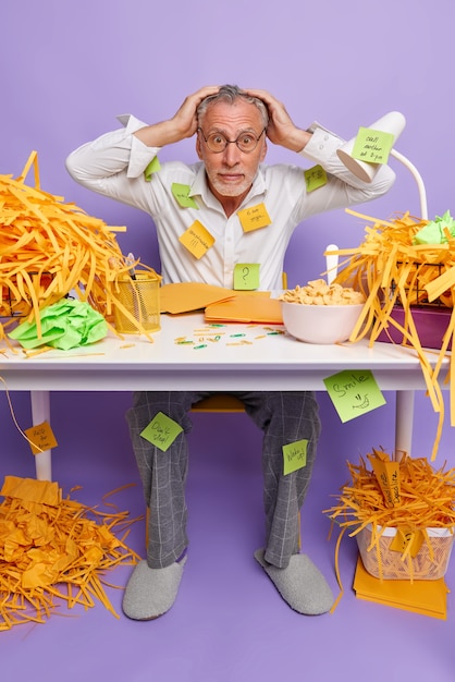 Terrified multitasking elderly man looks with panic keeps hands on head cannot decide what to do first embarrassed by deadline poses in coworking space wears formal apparel