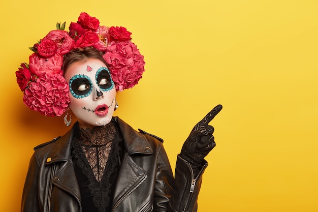 Terrified woman wears professional makeup for horror, dressed in black clothes, points away, wears gloves, red peonies wreath, celebrates Halloween holiday or Day of Death. Image of Calavera Catrina