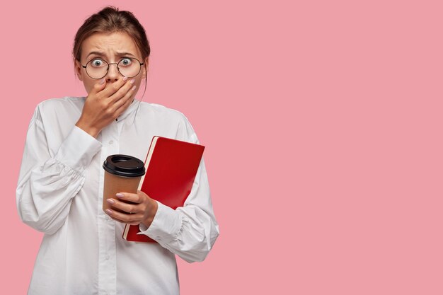 Terrified young woman with glasses posing against the pink wall