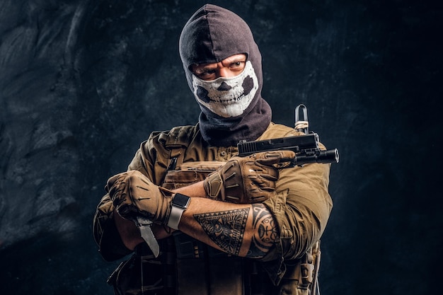 A terrorist in a military uniform and a skull balaclava holding a pistol and a knife and looks at the camera with a menacing look. Studio photo against a dark textured wall