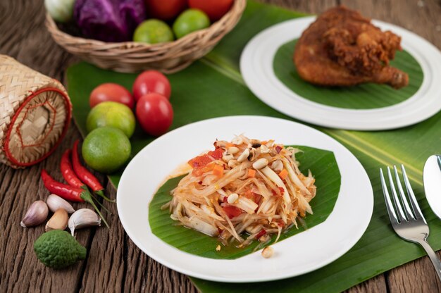 Thai papaya salad in a white plate on banana leaves with lime, tomatoes, eggplant, chili, garlic, peppers, salad, and peanuts.