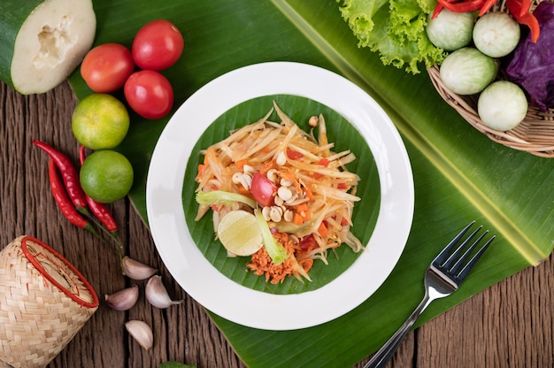 Thai papaya salad in a white plate on banana leaves with lime, tomatoes, eggplant, chili, garlic, peppers, salad, and peanuts.