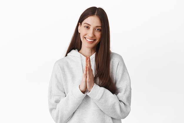 Thank you namaste smiling young woman look caring and pleased holding hands in beg pray gesture being grateful appreciate help standing over white background