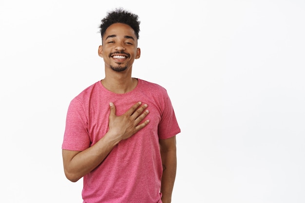 Thank you. Smiling african american man looking proud and happy, pleased by smth, holding hand on heart with gratitude, introduce himself, standing in pink t-shirt on white