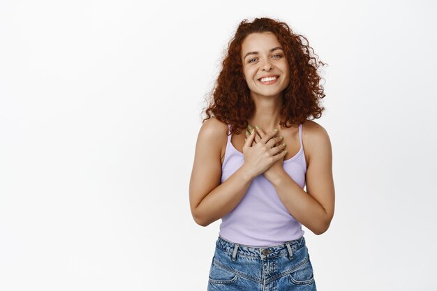 Thankful smiling redhead woman with curly hair, holding hands near chest and looking grateful, flattered with compliment, pleased of something good, white background.