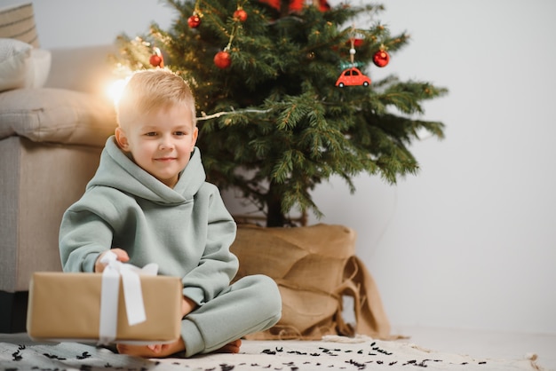 소년은 선물을 들고 크리스마스 트리 근처에 앉는다. 크리스마스 휴가를 위한 아이들. 프리미엄 사진