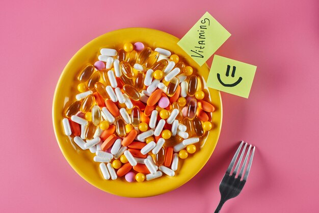 적절한 영양과 의학의 개념. 분홍색 배경에 노란색 접시에 캡슐, 알약 및 정제의 식이 보조제. 스티커에 비문 비타민