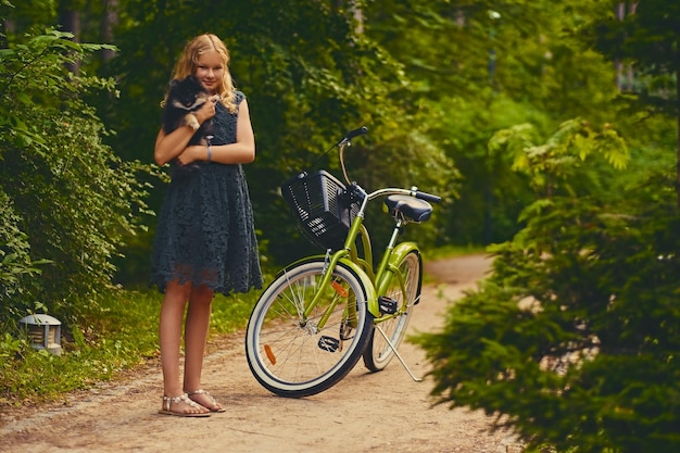 無料写真 ブロンドの女の子の全身画像は、自転車の背景の上にスピッツ犬を保持しています。