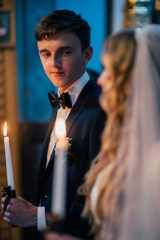 Жених держит свадебную свечу с коричневым бантом. горящая свеча. духовная пара, невеста. держит свечи во время свадебной церемонии в христианской церкви. закрыть счастливый жених смотрит на невесту.