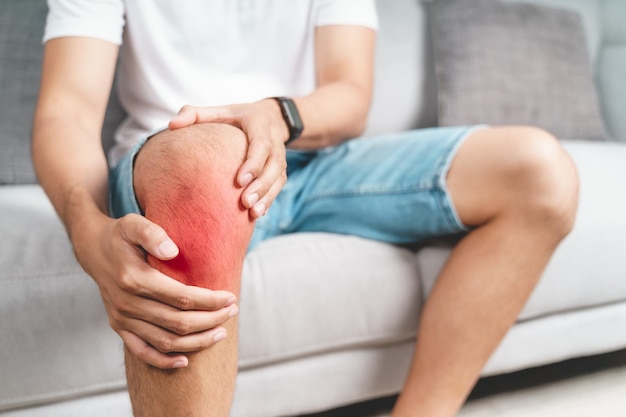 무릎 통증으로 고통받는 남자는 소파에 앉아 고통스러운 무릎을 잡고 마사지합니다.