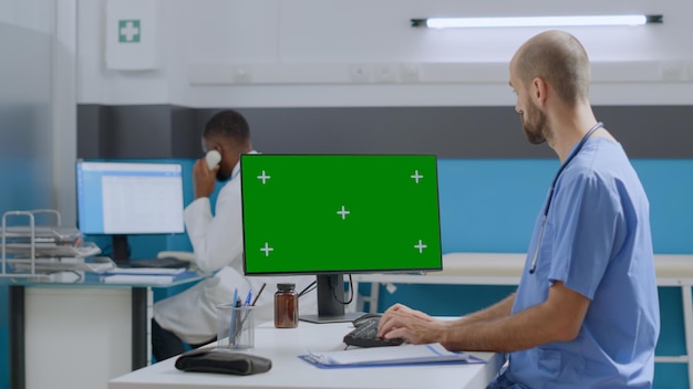 Therapist assistant man sitting at desk analyzing sickness report typing medical expertise on mock up greeen screen chroma key computer with isolated display in hospital office. Medicine concept