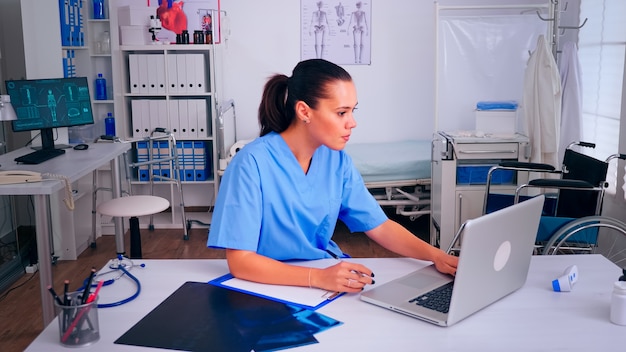 Therapist doctor assistant in uniform writing on clipboard, checking list of patients working on laptop in hospital clinic. Noting consulted patients online, making research, useful information