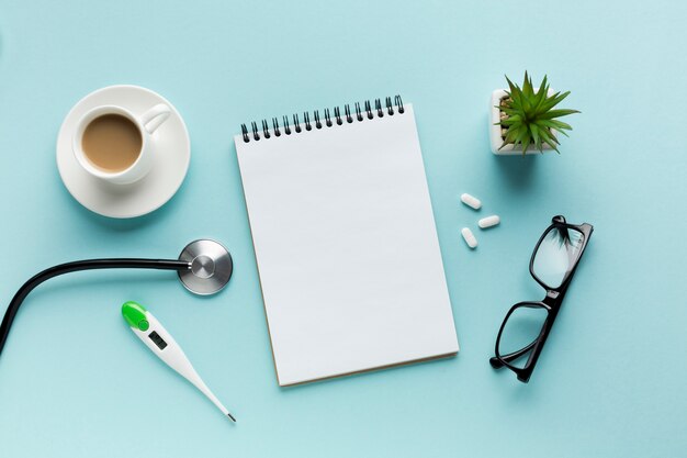 Thermometer; coffee cup; stethoscope with spiral notepad; pills and spectacles over blue surface