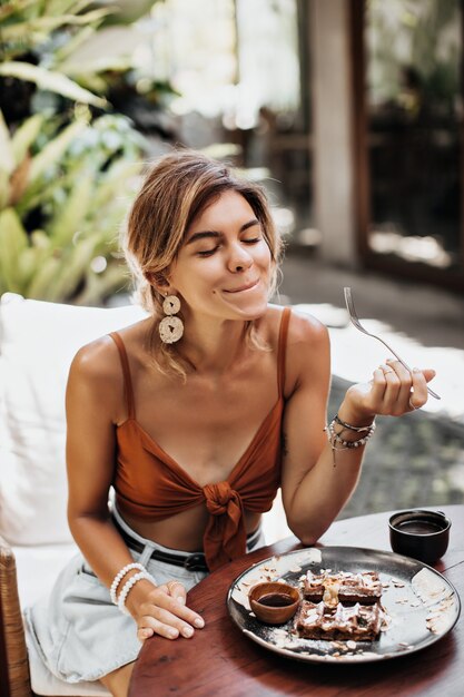 Thin tanned woman in brown bra and stylish denim shorts enjoys taste of waffle with cream, peanuts and maple syrup