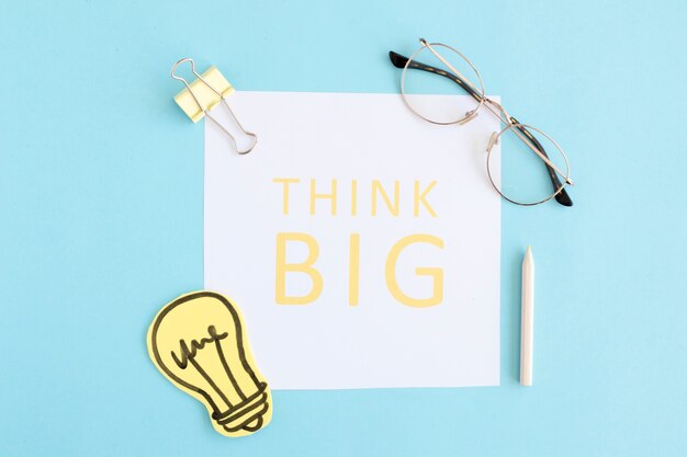 Think big text on white paper with light bulb, eyeglasses, bulldog pin and colored pencil on blue background