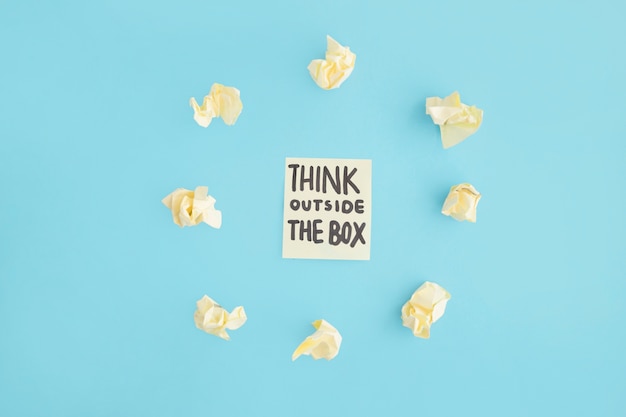 Think outside the box text on adhesive note surrounded with yellow crumpled paper over the blue backdrop