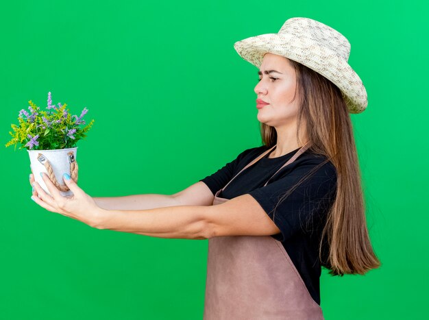 Thinking beautiful gardener girl in uniform wearing gardening hat holding out flower in flowerpot at side isolated on green background