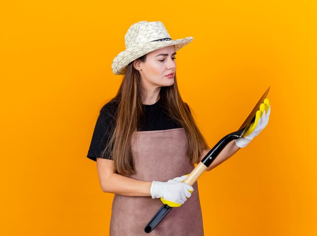 Думающая красивая девушка-садовник в униформе и садовой шляпе с перчатками держит и смотрит на лопату, изолированную на оранжевом фоне