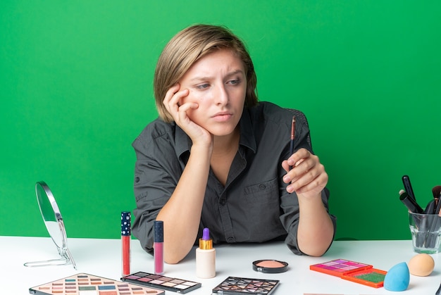 Foto gratuita pensare che una bella donna si siede al tavolo con gli strumenti per il trucco che tengono e guardano i pennelli per il trucco