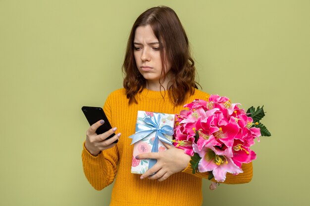 Thinking beautiful young girl on happy woman's day holding presents looking at phone in her hand isolated on olive green wall