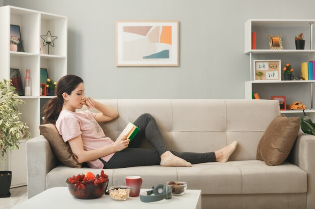 Thinking bites nails young girl lying on sofa behind coffee table reading book in living room