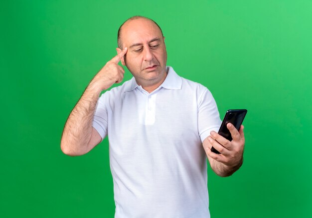 Thinking casual mature man holding and looking at phone putting finger on temple isolated on green wall