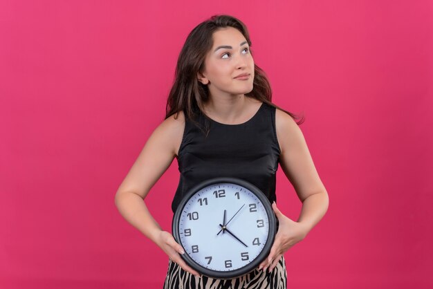 Thinking caucasian girl wearing black undershirt holding a wall clock on pink background