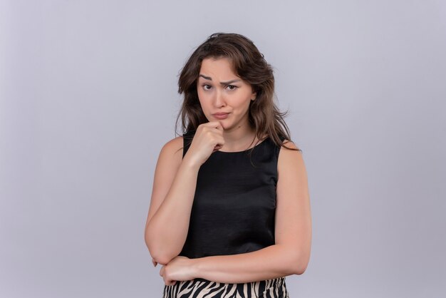 Thinking caucasian young girl wearing black undershirt grabbed jaw on white background