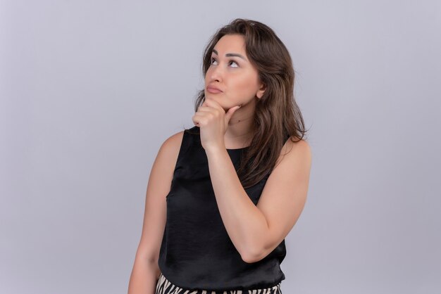 Thinking caucasian young girl wearing black undershirt put her hand on jaw on white background