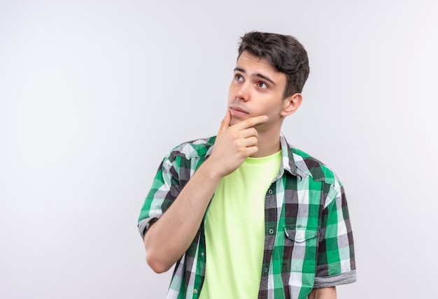 Thinking caucasian young guy wearing green shirt put his hand on chin on isolated white