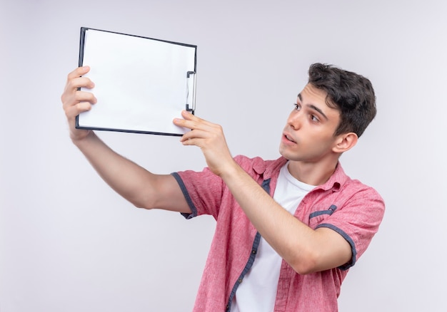 Thinking caucasian young guy wearing pink shirt raised clipboard on isolated white