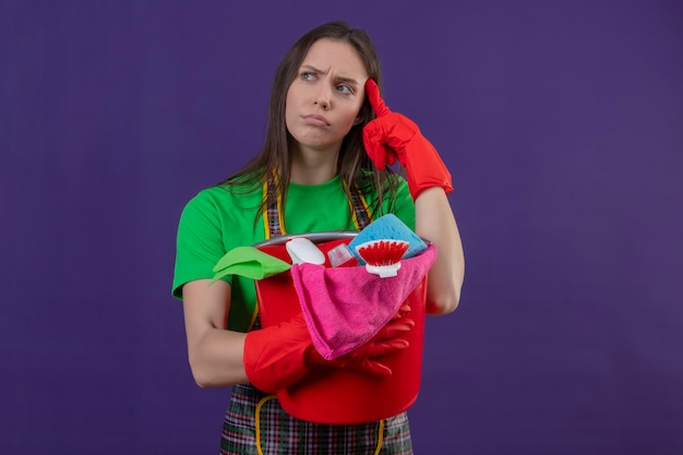 クリーニングツールを保持している赤い手袋で制服を着ている若い女の子を掃除することを考えて孤立した紫色の壁の額に彼女の指を置きます